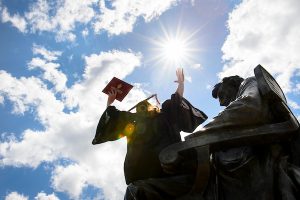 Wearing a graduation cap and gown, a soon-to-be-graduate holds a diploma cover overhead as she sits in silhouette in the lap of the Abraham Lincoln statue in front of Bascom Hall.