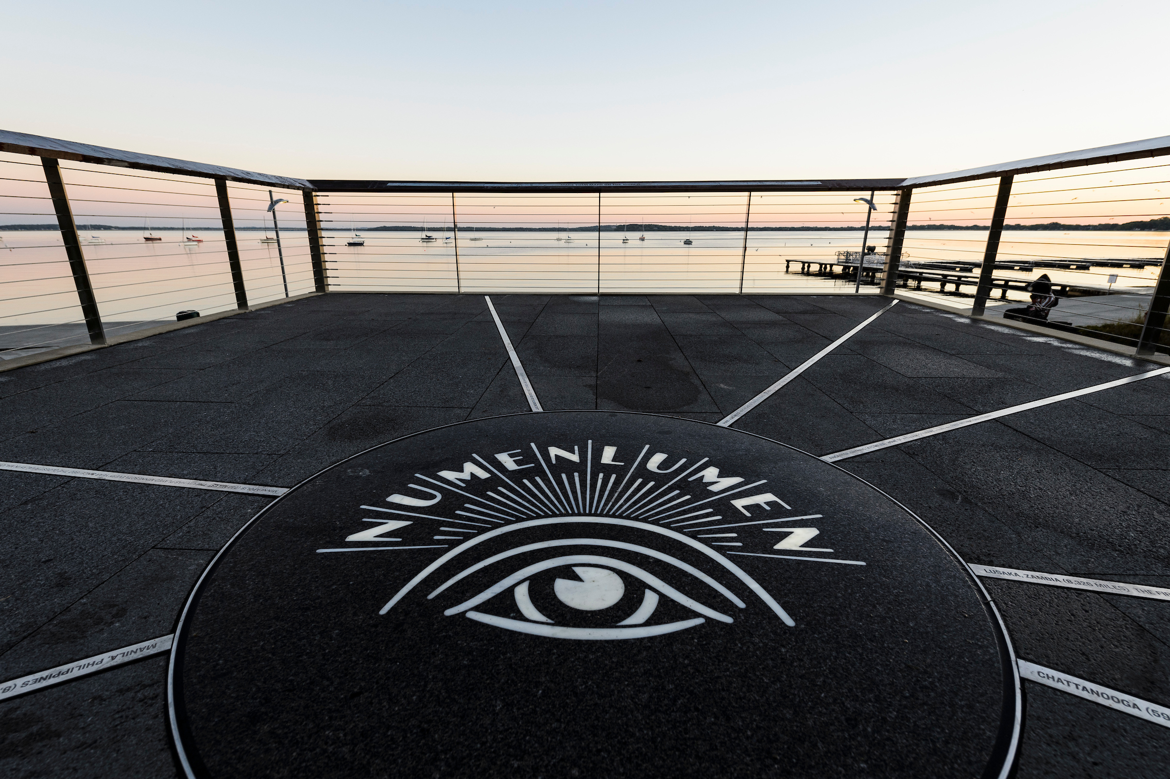 A backlit logo of the university's Numen Lumen seal is pictured in a dawn view of Lake Mendota from Alumni Park's Progress Point.