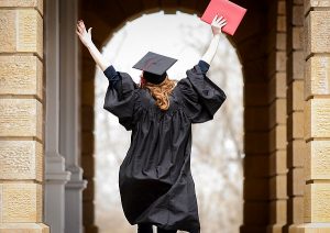 Wearing a graduation cap and gown, a soon-to-be-graduate holds a diploma cover overhead while walking in silhouette through the arched portico of Bascom Hall. 