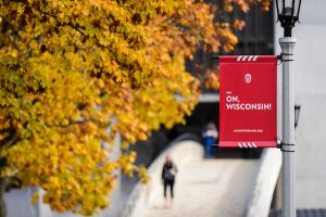 A red and white banner featuring the phrase "On, Wisconsin!" is pictured as a pedestrian walks down Bascom Hill during autumn