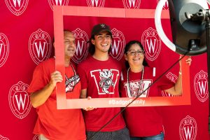 A Badger family stands in front of a red photo background and smiles for the camera in a photo booth during Family Weekend 2021.