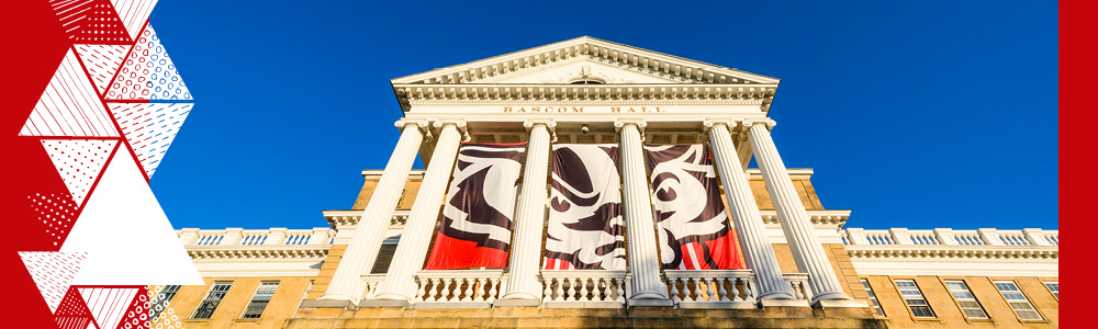 An up-close view of Bascom Hall on a sunny day with Bucky Badger flags between the columns of the hall.