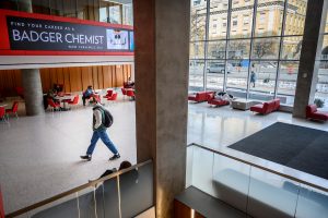 A student walks past red lounge furniture and a wall of windows in the Chemistry building lobby.