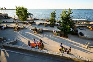 Students and community members seated around green, orange and yellow tables at the Memorial Union terrace in summer, Lake Mendota in the background.