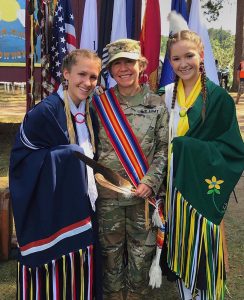 Three women stand in front of Native American and American flags, the two daughters wearing Ojibwe dress, the mom in military dress and a Native American sash.