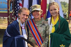 Three women stand in front of Native American and American flags, the two daughters wearing Ojibwe dress, the mom in military dress and a Native American sash.
