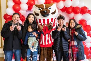 UW–Madison mascot Bucky Badger poses for a photo with a UW student and her extended family in front of a white and red balloon arch during Family Weekend.