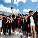 A row of graduates in caps and gowns smile at the camera on the field of Camp Randall Stadium as they jump and raise their hands in celebration.