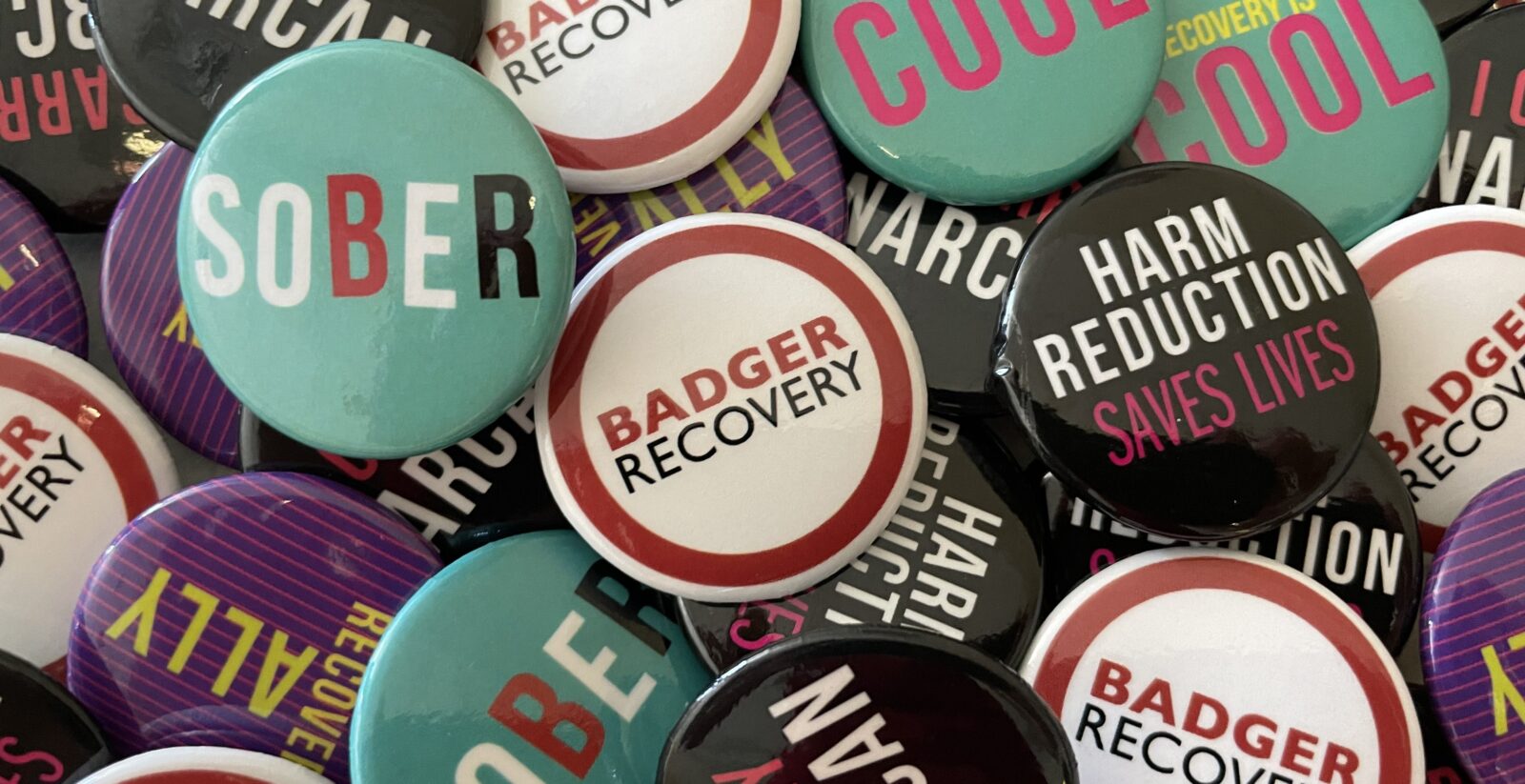 A collection of enamel pins that say Sober, Badger Recovery, Harm Reduction Saves lives, Recovery is Cool etc.